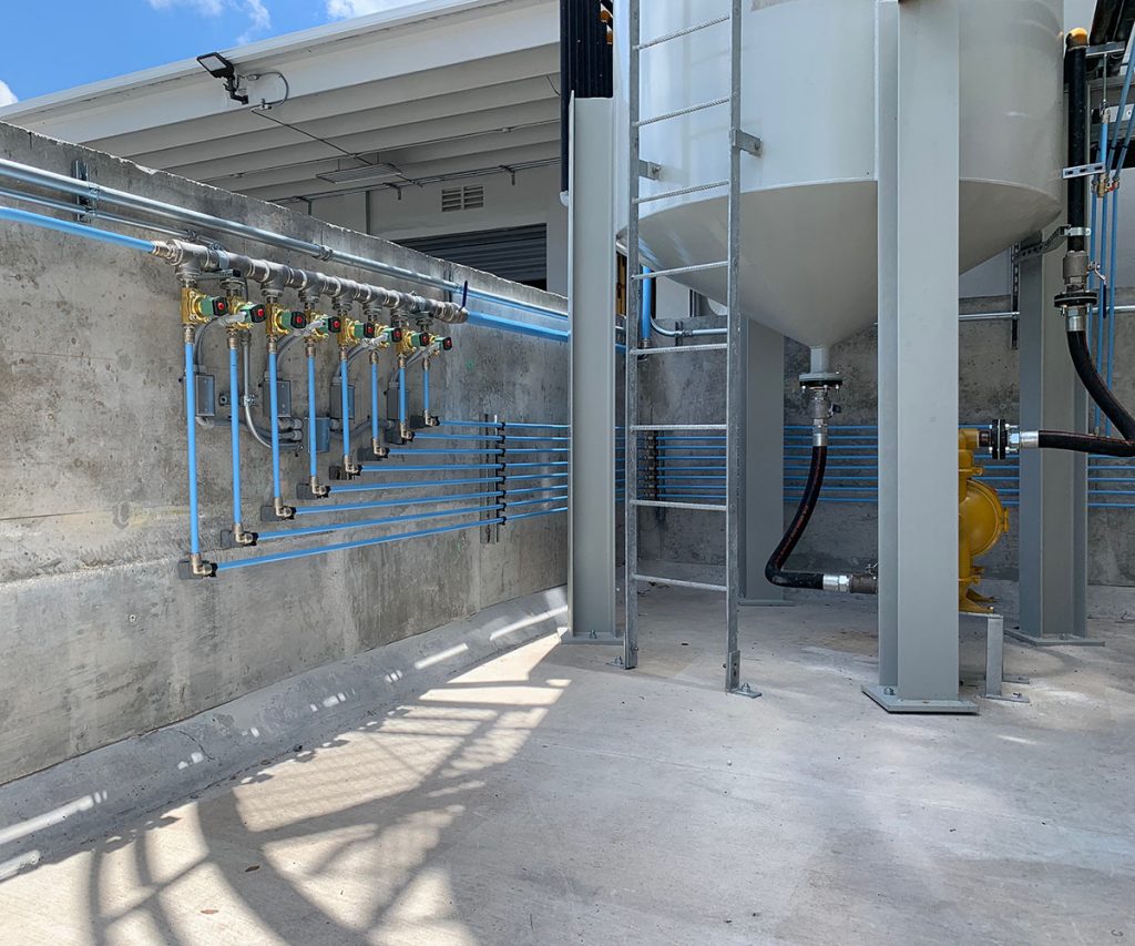 Industrial setting showing a section of a water treatment facility with pipework and structural supports, under bright sunlight casting sharp shadows, featuring Air Centers of Florida.