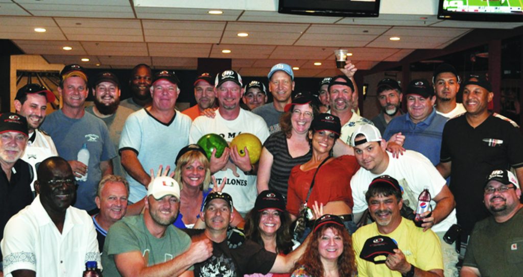 A group of cheerful people posing for a photo at a bowling alley, some holding bowling balls and wearing Air Centers of Florida team caps, ready for a fun game.