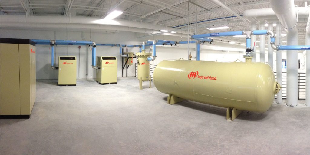 A spacious industrial room, part of Air Centers of Florida, featuring multiple air compressors and a large air tank, all connected by a network of overhead pipes, with a clean and organized layout.