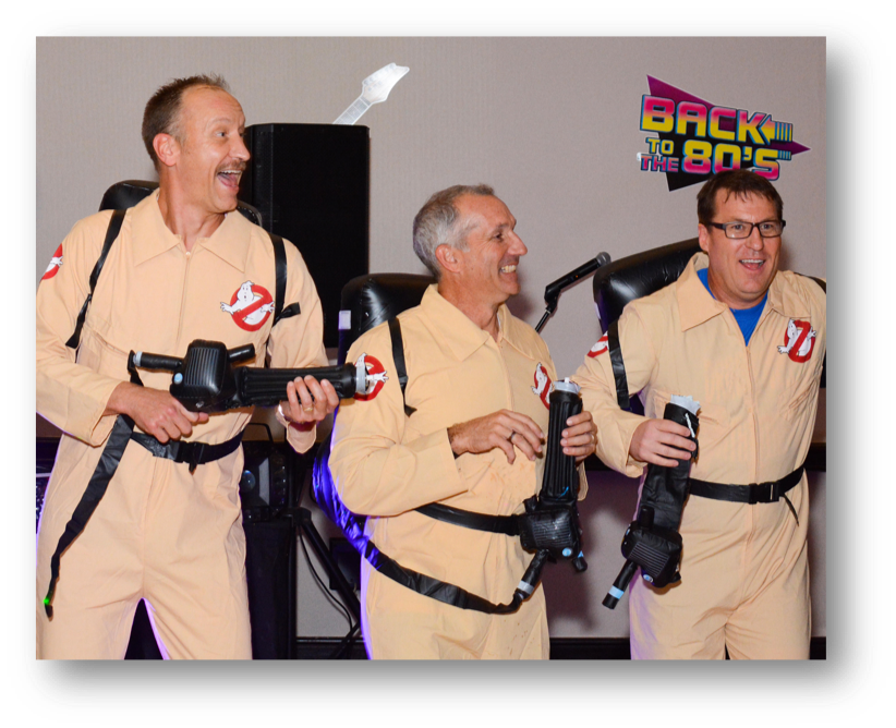 Three joyful individuals dressed as ghostbusters at an 80s-themed event, sponsored by Air Centers of Florida, complete with proton packs and the iconic logo, ready to bust some ghosts.