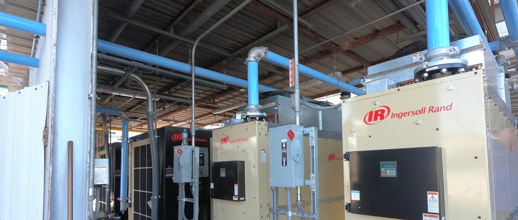 Industrial air compressors from Air Centers of Florida installed in a plant room with blue piping systems.