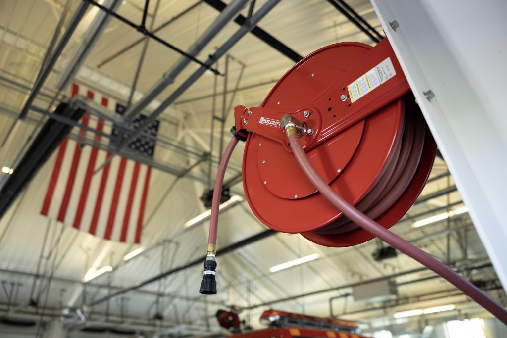 A red retractable hose reel mounted on the wall in a clean industrial or workshop environment at Air Centers of Florida, with an American flag in the background.